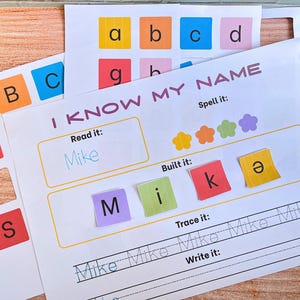 May include: A white educational worksheet titled I KNOW MY NAME with sections to read, build, trace, and write the name Mike. The sheet includes colorful square letter tiles and flower icons, with additional letter cutouts visible in the background.