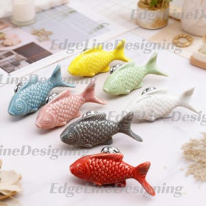 May include: A collection of seven ceramic fish-shaped cabinet knobs in various colors including yellow, light green, blue, pink, white, gray, and red. Each knob features a detailed scale texture and a silver-toned metal mounting base for attachment.
