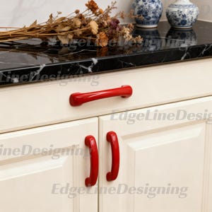 May include: Three glossy red cabinet pulls are mounted on off-white kitchen cabinetry. One horizontal pull is centred on the top drawer, while two vertical pulls are positioned on the lower cabinet doors, featuring a smooth, rounded design.