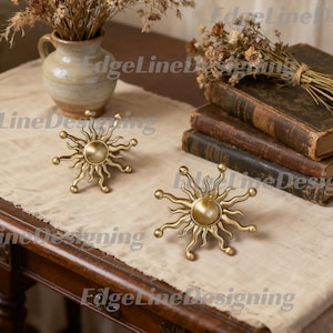 May include: Two gold-toned metal sun-shaped wall hooks or decorative knobs. Each piece features a central circular base surrounded by radiating, wavy, flame-like rays with rounded tips. The finish is a matte, brushed metallic gold texture.
