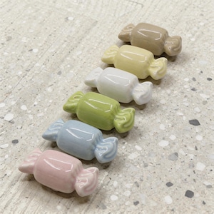 Könnte beinhalten: Eine Reihe von sechs keramischen, bonbonförmigen Figuren. Die Bonbons sind in Pastellfarben gehalten: Rosa, Hellblau, Grün, Weiß, Gelb und Beige. Jedes Bonbon hat ein gewickeltes Design mit gerüschten Kanten.