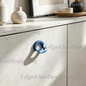 May include: A single blue cabinet knob with an irregular, organic, rounded shape attached to a light-coloured, speckled stone-textured cabinet door. The knob features a smooth, matte finish and casts a distinct shadow on the cabinet surface.