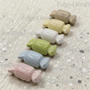 Op de afbeelding: Een set van zes keramische ornamenten in de vorm van snoepjes, gerangschikt in een diagonale lijn. De kleuren zijn beige, lichtgeel, wit, lichtgroen, lichtblauw en zachtroze. Elk stuk heeft een gladde, glanzende afwerking en gedraaide uiteinden.