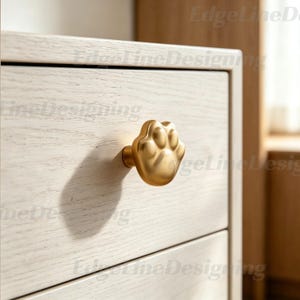 Puede incluir: Un pomo dorado con forma de huella de pata está fijado a un cajón de madera de color claro. El pomo es un diseño tridimensional detallado, que proyecta una sombra sobre el cajón. El cajón forma parte de un mueble más grande.