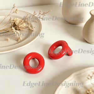 May include: A pair of bright red earrings featuring a smooth, glossy, circular donut-shaped design. Each earring has a hollow center and a polished finish, creating a bold and vibrant look. The earrings are presented as a set of two matching pieces.