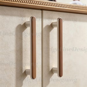 Puede incluir: Primer plano de dos tiradores de puerta de armario. Cada tirador tiene una base plateada clara con un centro vertical rectangular de color madera marrón. Las puertas del armario son de color beige claro. La parte superior del armario tiene una moldura decorativa tallada en marrón.