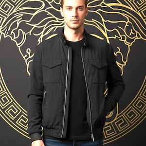 May include: A man wearing a black bomber jacket with a silver zipper, a black t-shirt, and dark blue jeans. The background is black with a gold decorative pattern.