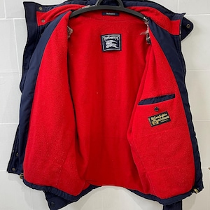 Peut inclure: Veste bleu marine avec intérieur rouge, suspendue à un cintre noir. La veste présente une étiquette Burberry et une petite poche avec une étiquette verte et dorée. La veste est ouverte, révélant la doublure rouge.