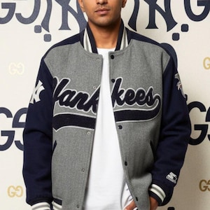 May include: A gray and navy blue Yankees letterman jacket with the word "Yankees" in white script across the chest. The jacket has a white collar and cuffs, and is worn over a white t-shirt. The background features a repeating pattern of the Yankees logo.