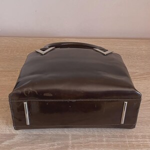 May include: A brown, glossy handbag with a rectangular shape. The bag features a curved handle and silver-colored metal accents at the handle's base and on the front. The bag is sitting on a light-colored wooden surface.