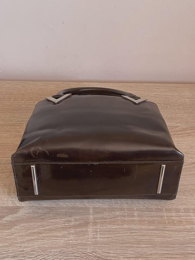 May include: A brown, glossy handbag with a rectangular shape. The bag features a curved handle and silver-colored metal accents at the handle's base and on the front. The bag is sitting on a light-colored wooden surface.