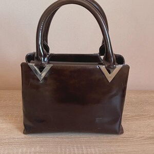 May include: A brown handbag with a glossy finish and two curved handles. The bag has a rectangular shape with silver-colored V-shaped accents on the front. The bag is sitting on a wooden surface.