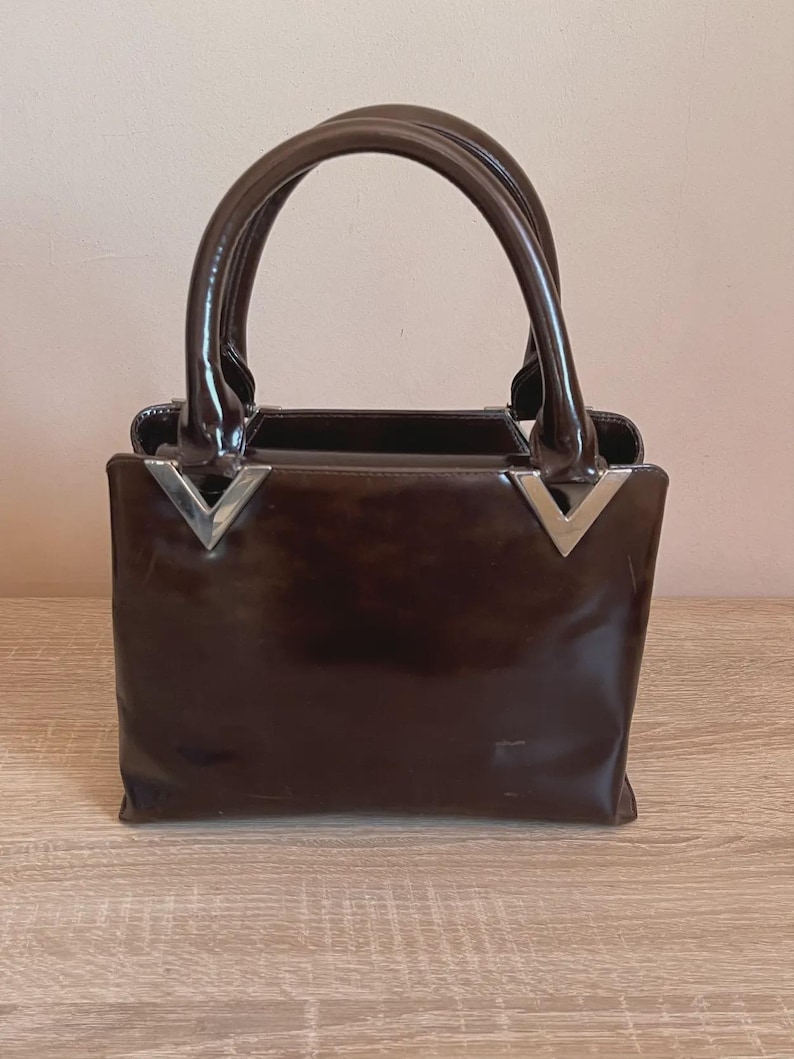 May include: A brown handbag with a glossy finish and two curved handles. The bag has a rectangular shape with silver-colored V-shaped accents on the front. The bag is sitting on a wooden surface.