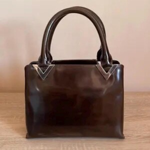 May include: A glossy, dark brown handbag with two rounded handles. The bag has a rectangular shape with silver-colored metal accents at the top corners. The bag is sitting on a light-colored wooden surface.