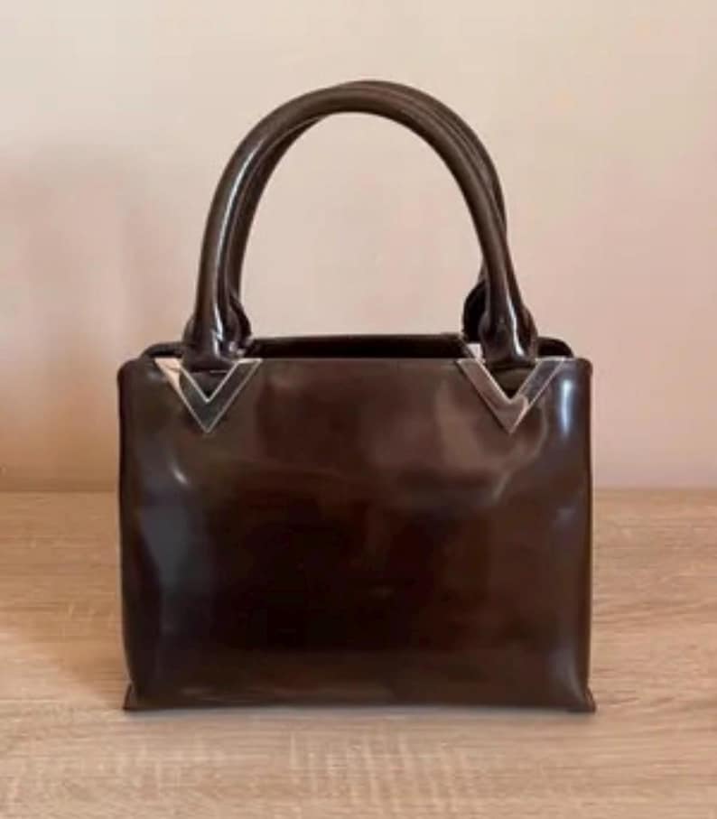 May include: A glossy, dark brown handbag with two rounded handles. The bag has a rectangular shape with silver-colored metal accents at the top corners. The bag is sitting on a light-colored wooden surface.