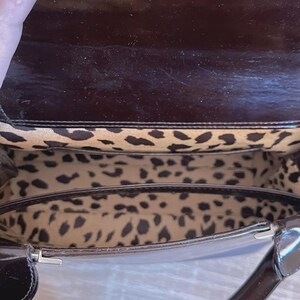 May include: A dark brown, patent leather handbag with a glossy finish. The interior features a leopard print lining. The bag has a handle and a silver-colored clasp.