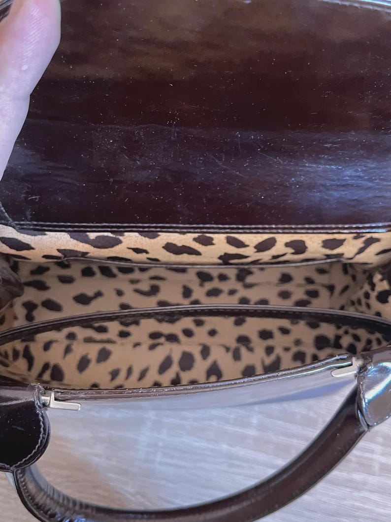 May include: A dark brown, patent leather handbag with a glossy finish. The interior features a leopard print lining. The bag has a handle and a silver-colored clasp.
