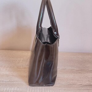 May include: A glossy, dark brown handbag with a structured design. The bag features a rectangular shape with a rounded top and two handles. The material appears to be patent leather or a similar shiny material, reflecting light. The bag is set against a neutral background.