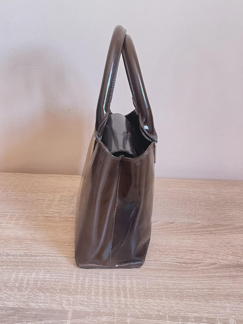 May include: A glossy, dark brown handbag with a structured design. The bag features a rectangular shape with a rounded top and two handles. The material appears to be patent leather or a similar shiny material, reflecting light. The bag is set against a neutral background.