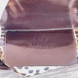 May include: A brown Valentino Couture handbag with a glossy finish. The bag features a flap with the brand's logo embossed on it. The interior lining has a leopard print pattern.