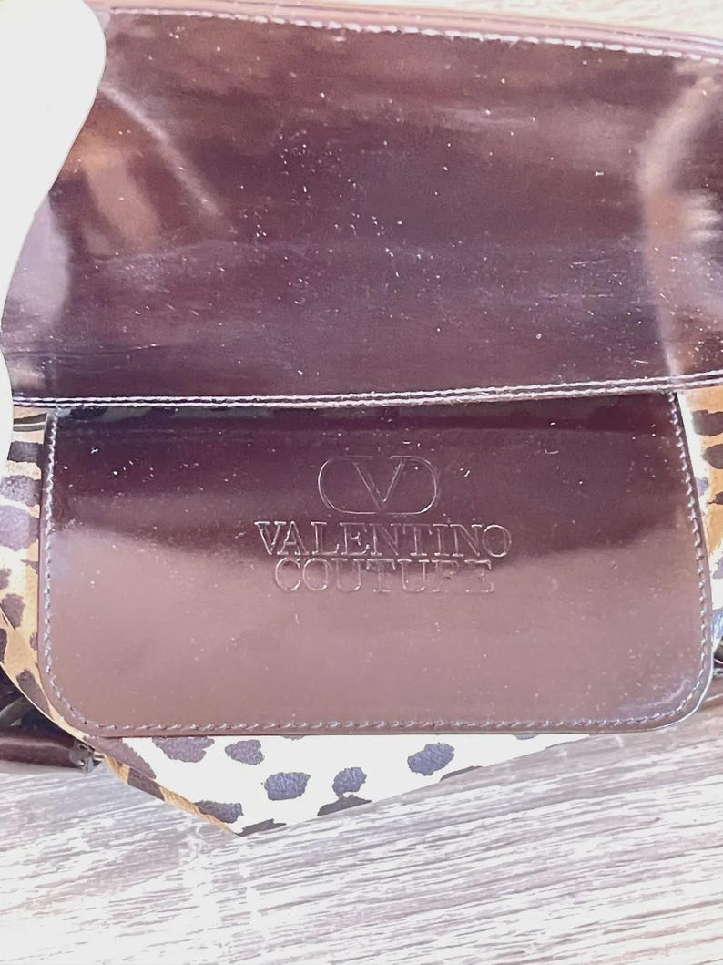 May include: A brown Valentino Couture handbag with a glossy finish. The bag features a flap with the brand's logo embossed on it. The interior lining has a leopard print pattern.