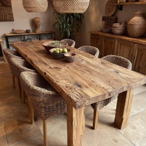 May include: A large, rectangular wooden dining table with a natural finish, accompanied by woven wicker chairs. The table has thick, sturdy legs. Bowls and plates are on the table. Woven pendant lights hang above the table, creating a warm atmosphere. The table is approximately 240 cm long.