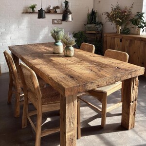 Op de afbeelding: Een rechthoekige houten eettafel met een natuurlijke afwerking en dikke poten. Stoelen met geweven rugleuningen omringen de tafel. Twee vazen met gedroogde bloemen staan op de tafel. De kamer heeft een warme, uitnodigende sfeer.