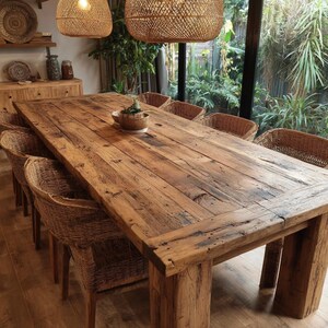 May include: A large, rectangular wooden dining table with a rustic, weathered appearance. The table is surrounded by woven wicker chairs. Two woven pendant lights hang above the table. A small bowl with a plant sits on the table's surface.