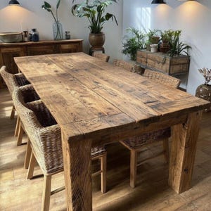 Op de afbeelding: Een grote, rechthoekige houten eettafel met een rustieke, verweerde uitstraling. De tafel is omringd door stoelen van geweven riet. Het hout heeft een natuurlijke, warme bruine tint en de tafel is stevig.