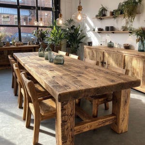 Op de afbeelding: Een grote, rustieke houten eettafel met bijpassende stoelen. De tafel is gemaakt van gerecycled hout en heeft een natuurlijke, verweerde uitstraling. De stoelen zijn ook van hout, met een eenvoudig, klassiek ontwerp. De kamer is ingericht met planten en glazen vazen.
