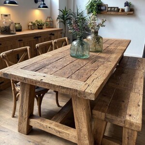 Op de afbeelding: Een rustieke houten eettafel met bijpassende bank. De tafel heeft een dik, rechthoekig blad van gerecyclede houten planken. De bank heeft een vergelijkbaar ontwerp. Twee glazen vazen met bloemen staan op de tafel. De tafel is omringd door houten stoelen.
