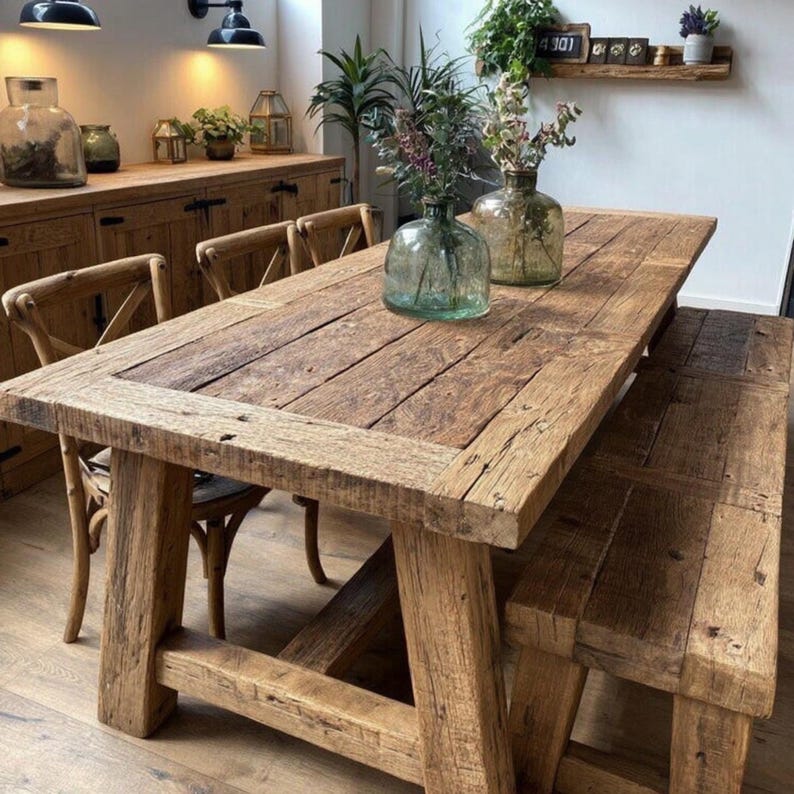 Op de afbeelding: Een rustieke houten eettafel met bijpassende bank. De tafel heeft een dik, rechthoekig blad van gerecyclede houten planken. De bank heeft een vergelijkbaar ontwerp. Twee glazen vazen met bloemen staan op de tafel. De tafel is omringd door houten stoelen.