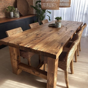 May include: A large, rustic wooden dining table with a natural finish. The table is supported by thick, square legs and is surrounded by woven wicker chairs. The table is approximately 244 cm long and 102 cm wide, perfect for a dining room.
