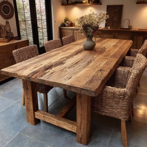 May include: A substantial, rectangular wooden dining table with a natural finish. The table is supported by thick, sturdy legs. Woven wicker chairs surround the table. A vase of dried flowers sits on the table's surface, creating a rustic dining space.