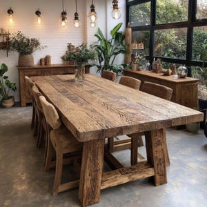 Op de afbeelding: Een grote, rustieke houten eettafel met een rechthoekig blad en dikke poten. Acht houten stoelen met gestoffeerde zittingen omringen de tafel. De kamer heeft een neutraal kleurenpalet, natuurlijk licht en decoratieve planten. Tafel ongeveer 240 cm lang.