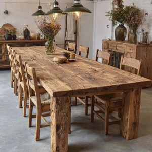Op de afbeelding: Een grote, rustieke houten eettafel met acht bijpassende stoelen. De tafel is gemaakt van gerecycled hout en heeft een natuurlijke afwerking. De stoelen hebben geweven zittingen. Boven de tafel hangen drie groene hanglampen. De kamer is gedecoreerd met droogbloemen.