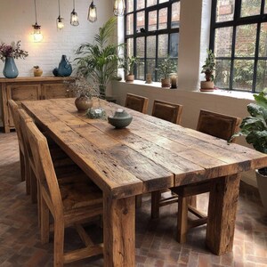 Op de afbeelding: Een grote, rustieke houten eettafel met bijpassende stoelen. De tafel is gemaakt van gerecycled hout, met de natuurlijke nerf en textuur. De kamer is ingericht met planten, vazen en hanglampen, wat een warme sfeer cre&euml;ert.