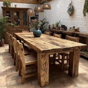 Op de afbeelding: Een rustieke houten eettafel met bijpassende stoelen. De tafel is gemaakt van dik, gerecycled hout en heeft een natuurlijke, verweerde afwerking. Stoelen met geweven zittingen omringen de tafel. Boven hangen drie geweven hanglampen.