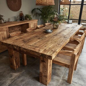 May include: A large, reclaimed wooden dining table with a rectangular top and chunky legs. Matching wooden chairs with woven seats are arranged around the table. The wood has a natural, aged look. A sideboard and decorative items are in the background.