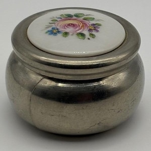 May include: A small, round, silver-toned jar with a lid. The lid is white ceramic and features a hand-painted floral design with pink, purple, and blue flowers. The jar's body has a slightly textured appearance.