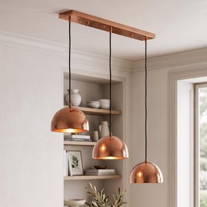 Könnte beinhalten: Eine dreiflammige Pendelleuchte mit gehämmerten kupferfarbenen Kuppelschirmen, die an schwarzen Kabeln von einem rechteckigen Deckenbaldachin aus Kupfer hängen. Die metallische Oberfläche reflektiert das Licht und erzeugt einen warmen Schein.