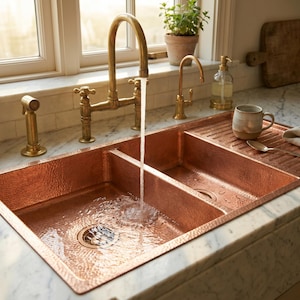 Peut inclure: Un évier de cuisine en cuivre à deux bacs et une égouttoir intégré. L'évier a une texture martelée et est associé à un robinet en laiton. Un filet d'eau coule dans l'évier. Une tasse, une cuillère et un torchon sont sur l'égouttoir.