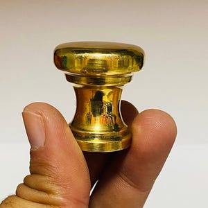 Puede incluir: Un tapón de botella decorativo de color dorado. Tiene una parte superior redondeada y una base ensanchada, con una sección media cilíndrica. El acabado metálico refleja la luz, resaltando su forma y diseño. Este artículo se utiliza para sellar botellas.