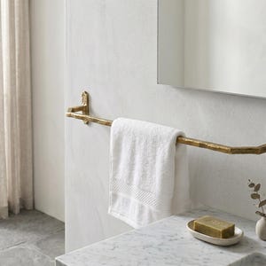 May include: A wall-mounted towel bar with a textured, hammered gold finish. The horizontal bar is secured by two rectangular mounting plates at each end. A plain white rectangular hand towel is draped over the center of the metal bar.