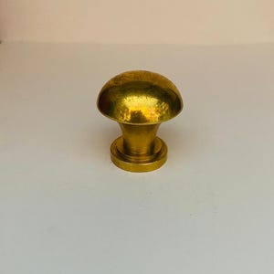 Peut inclure: Un bouton de meuble doré en forme de champignon. Le bouton a un dessus arrondi et une base cylindrique. La surface du bouton a une texture martelée, lui donnant un aspect rustique. Le bouton est conçu pour les tiroirs ou les armoires.