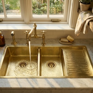 May include: A double-basin hammered brass kitchen sink with an integrated drainboard. A brass tap is running water into the sink. A brown glass soap dispenser and a brass sprayer are on the countertop. The sink is set against a white marble worktop.