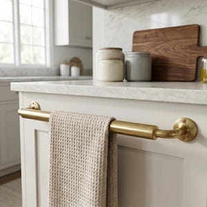 May include: A brushed brass towel bar mounted horizontally on a white cabinet door. A beige waffle-textured hand towel hangs over the center of the metal bar. The bar features circular mounting plates at each end, secured with visible screws.