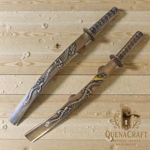 Puede incluir: Dos manijas de puerta decorativas de latón con forma de katanas japonesas. Las manijas presentan diseños de dragones y son de color plata y bronce. El texto "QUENACRAFT ANTIQUE BRASS HARDWARE" es visible.