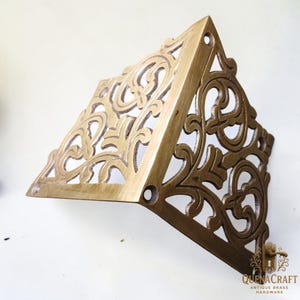 May include: A brass-colored, triangular corner bracket with ornate, swirling cut-out designs. The bracket has a vintage aesthetic and features pre-drilled holes for mounting. The product is from QuenaCraft Antique Brass Hardware.