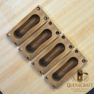 May include: Four antique brass recessed pulls, rectangular with an oval grip. Each has four screw holes. The pulls are a warm brown with a darker, aged finish in the recessed area. The logo "QUENACRAFT ANTIQUE BRASS HARDWARE" is in the bottom right corner.